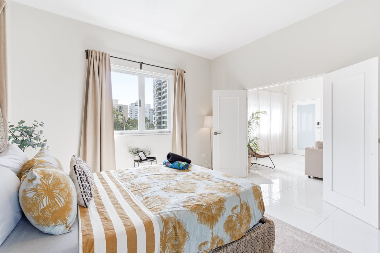 Furnished bedroom with a patterned bed and a large window overlooking nearby buildings.
