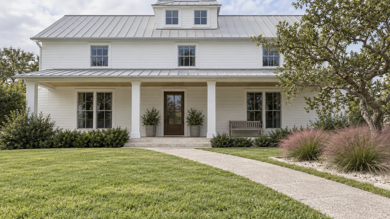 White timber-sided home with a metal roof, deep limestone porch, lawn and pink muhly grass beside a sculptural live oak.