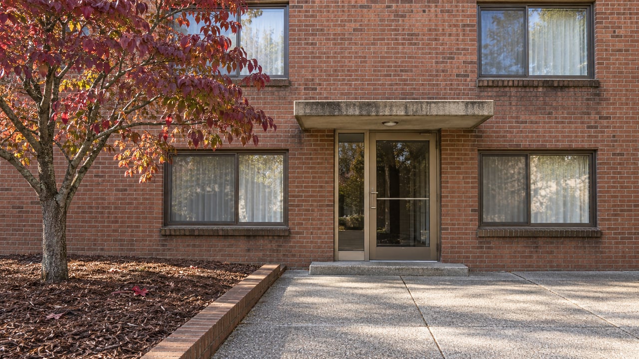 Established red-brick apartment entrance with bronze-framed windows, a concrete canopy, and an autumn dogwood beside the approach.