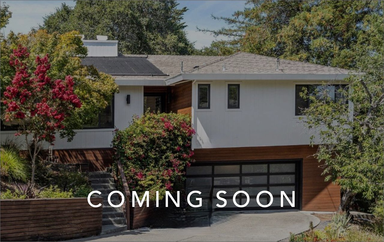 Modern white-and-wood home with flowering landscaping and “COMING SOON” lettering.