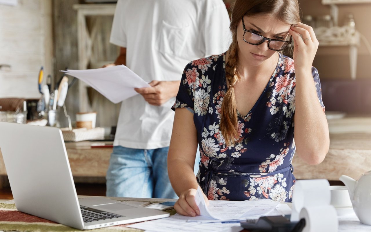 Worried woman reviewing paperwork at a kitchen counter, with a person holding papers behind her.