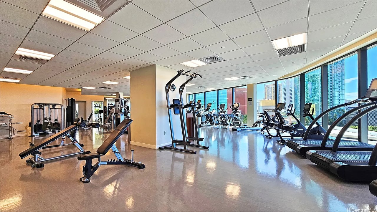 Condominium fitness center with treadmills and weight machines beside floor-to-ceiling windows overlooking the city.