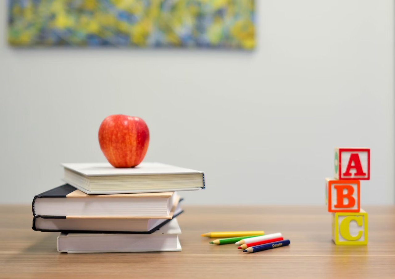 Red apple on stacked books beside colored pencils and ABC blocks.