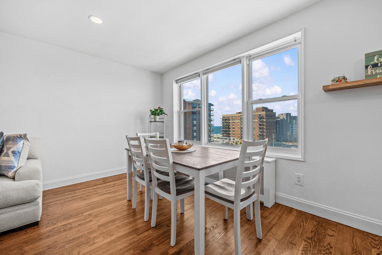 Dining area with white chairs, hardwood floors, and large windows overlooking city buildings and water.
