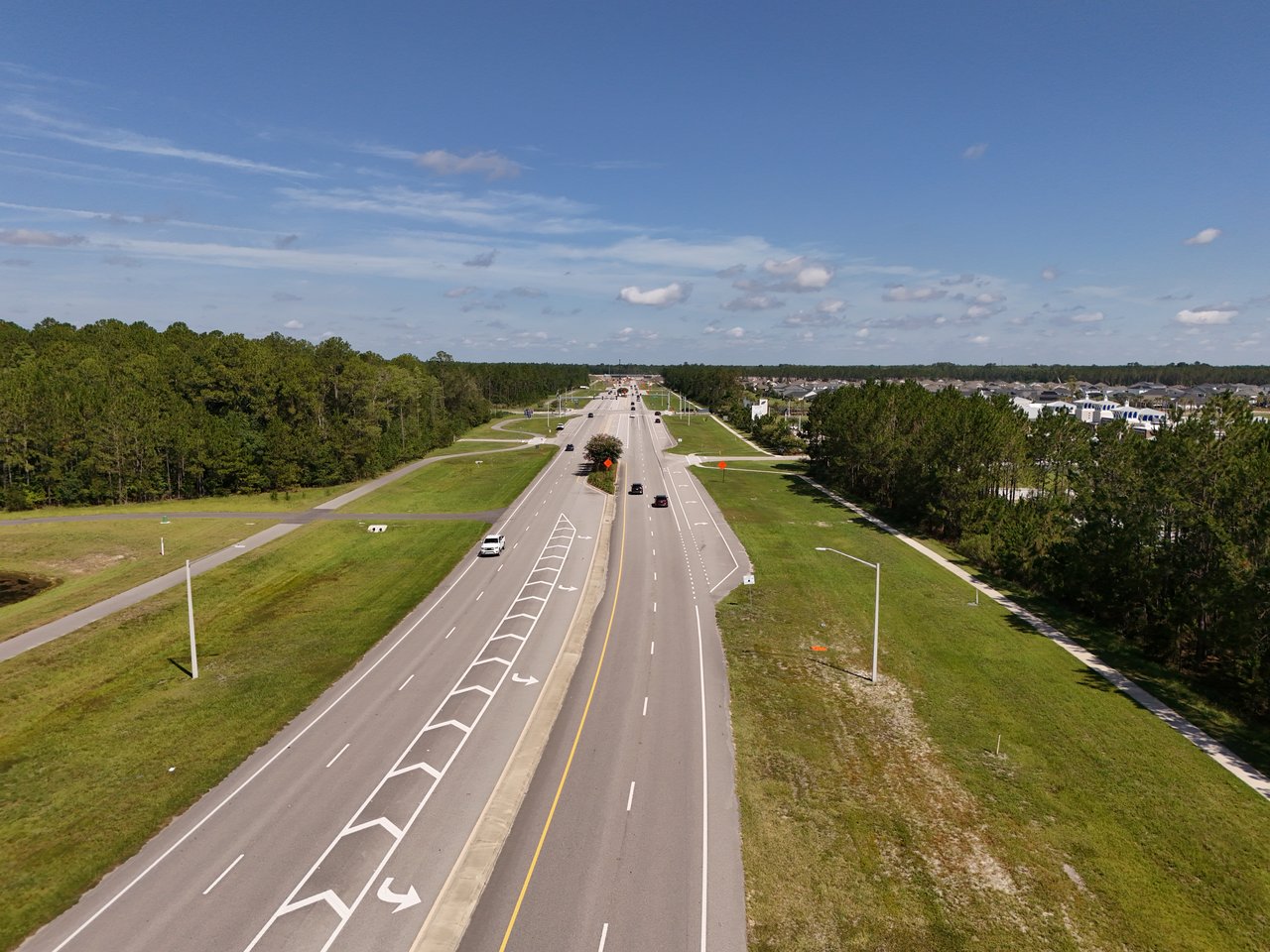 Aerial view of a divided road leading toward homes, bordered by grassy verges and woods.