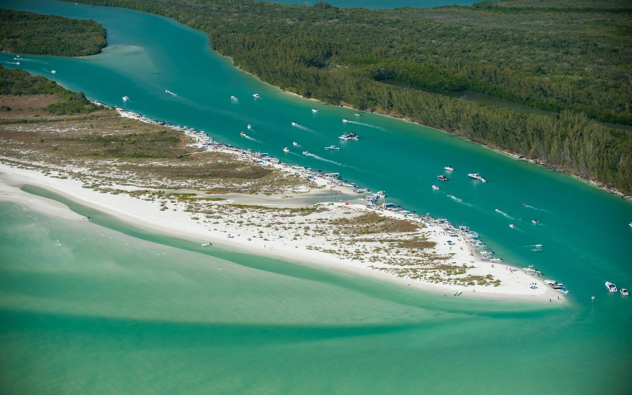 Aerial sandbar between turquoise waterways, lined with boats and backed by wooded shoreline.