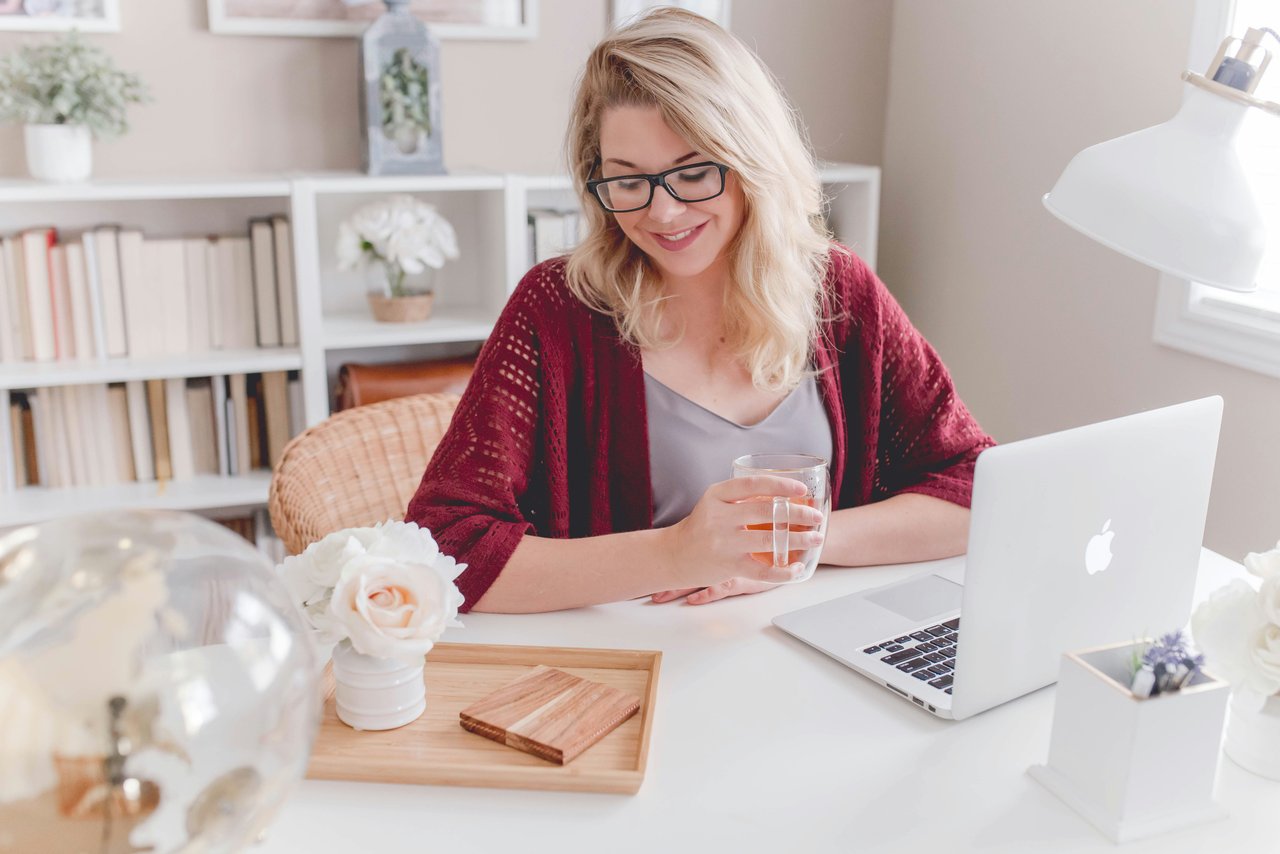 A woman sitting at a bright organized home office desk with a laptop and cup of tea, smiling calmly