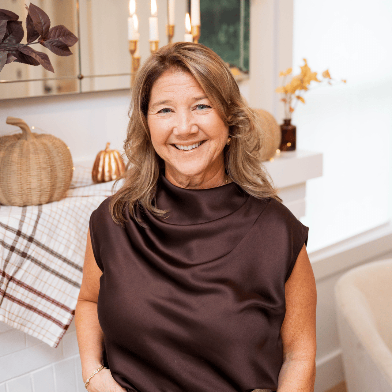 Smiling woman in a dark brown blouse beside a candlelit mantel with woven decor.