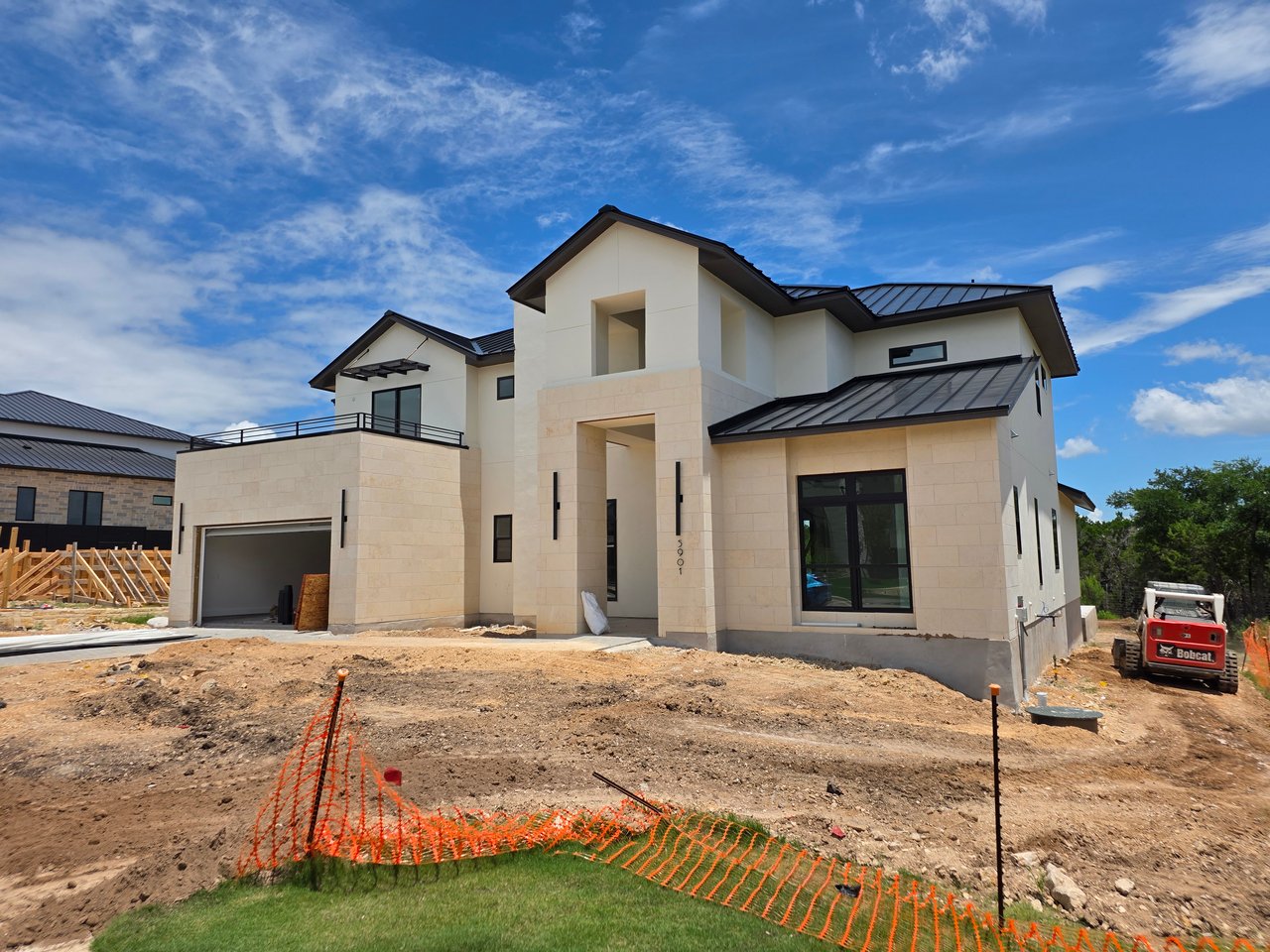 Modern two-story house under construction with pale stone walls, dark metal roofs, and orange safety fencing.