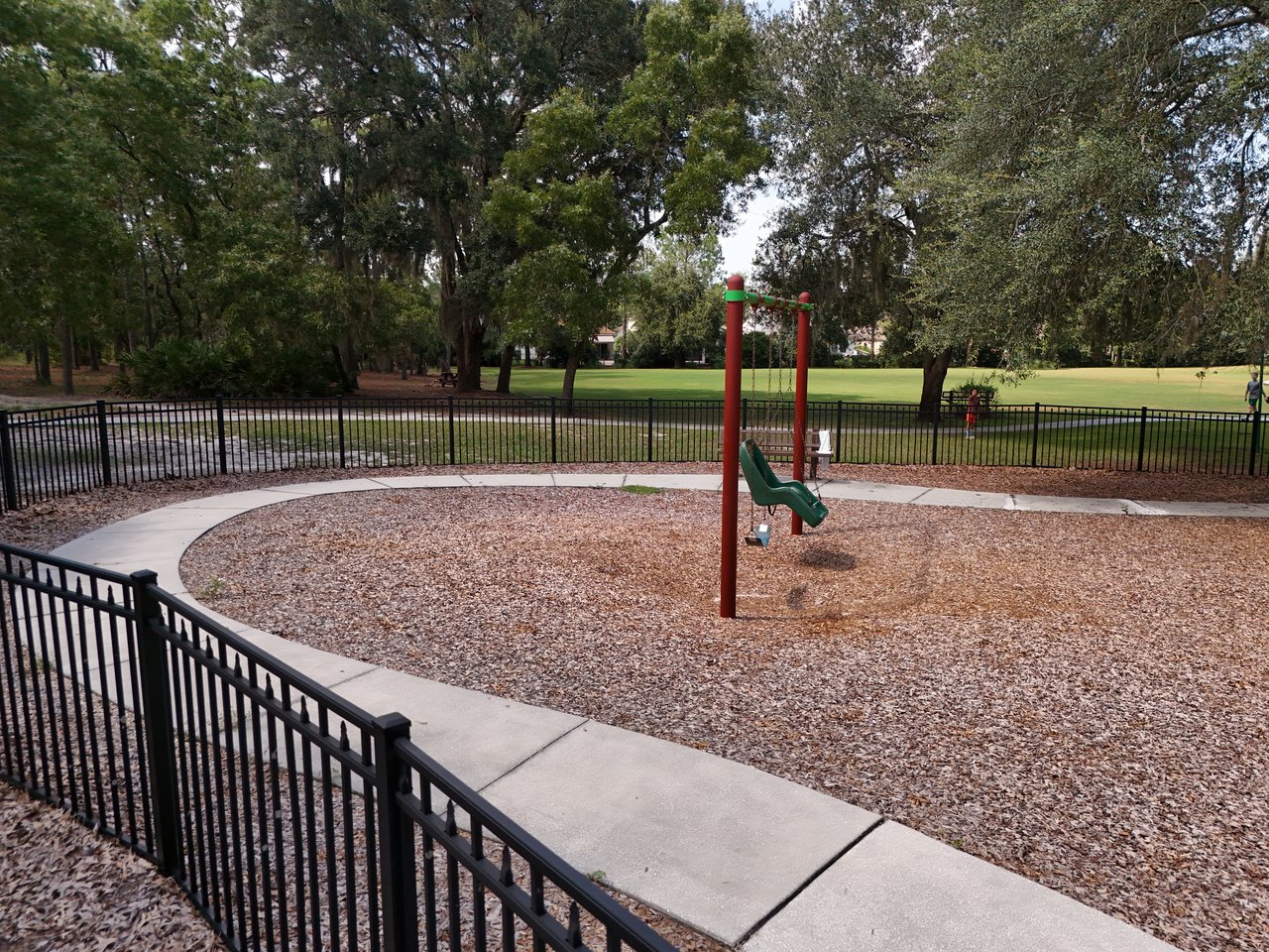 Fenced playground with a small green slide beneath mature trees.