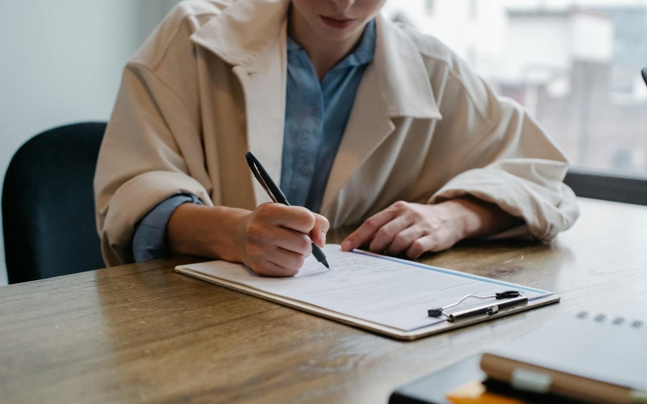 Person signing paperwork on a clipboard at a wooden table.