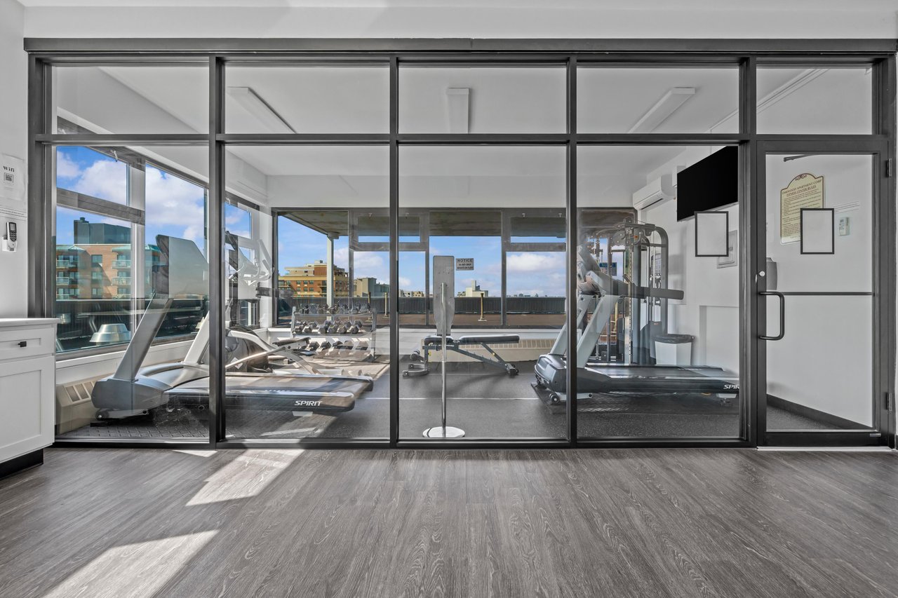 Shared fitness room with treadmills and weight equipment behind glass walls, overlooking nearby buildings.