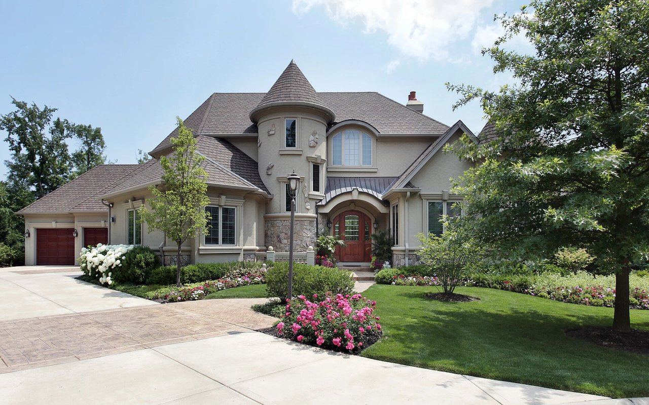 Large stucco home with a round turret, red garage doors, and colorful landscaped flower beds.