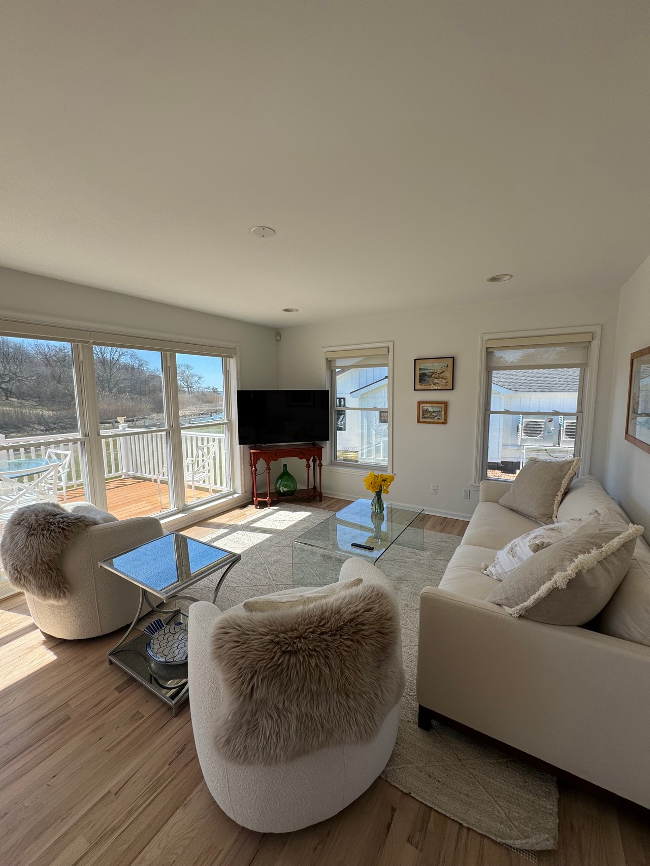 Furnished living room with cream seating and large windows overlooking a deck and glimpse of water.