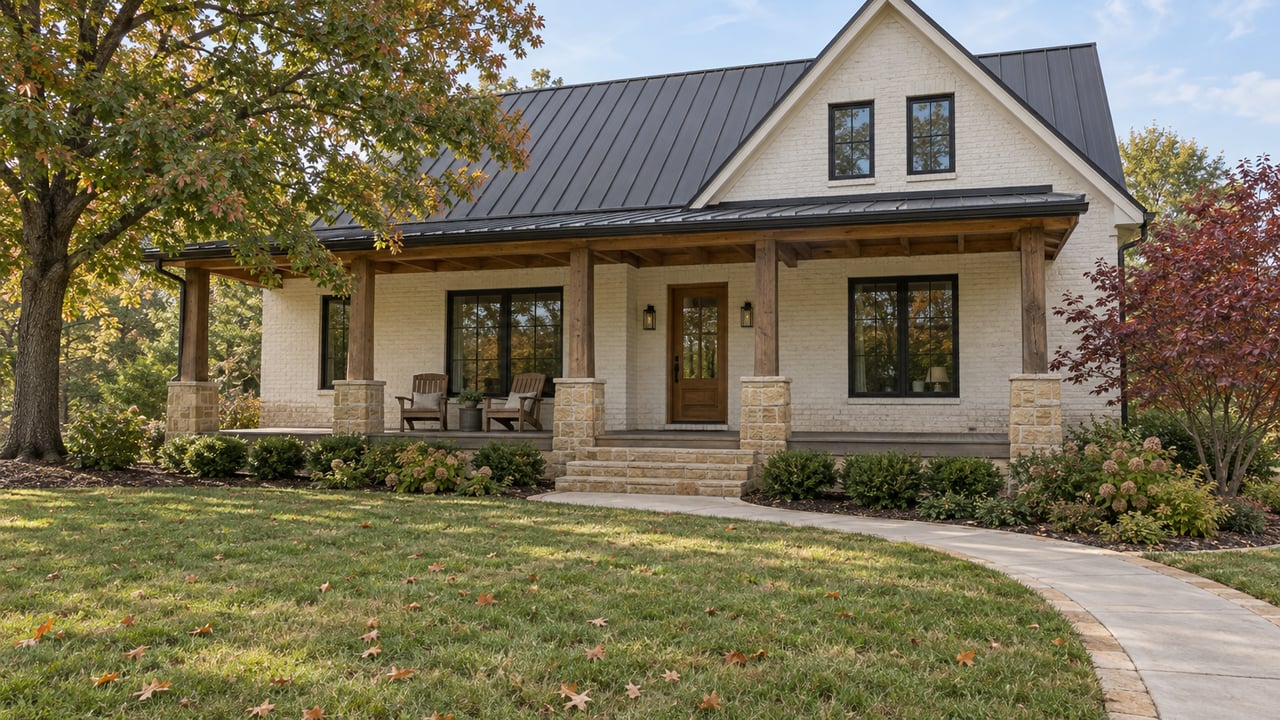 An ivory brick home with a timber porch and charcoal roof beyond a broad autumn lawn, framed by an oak and a burgundy dogwood.