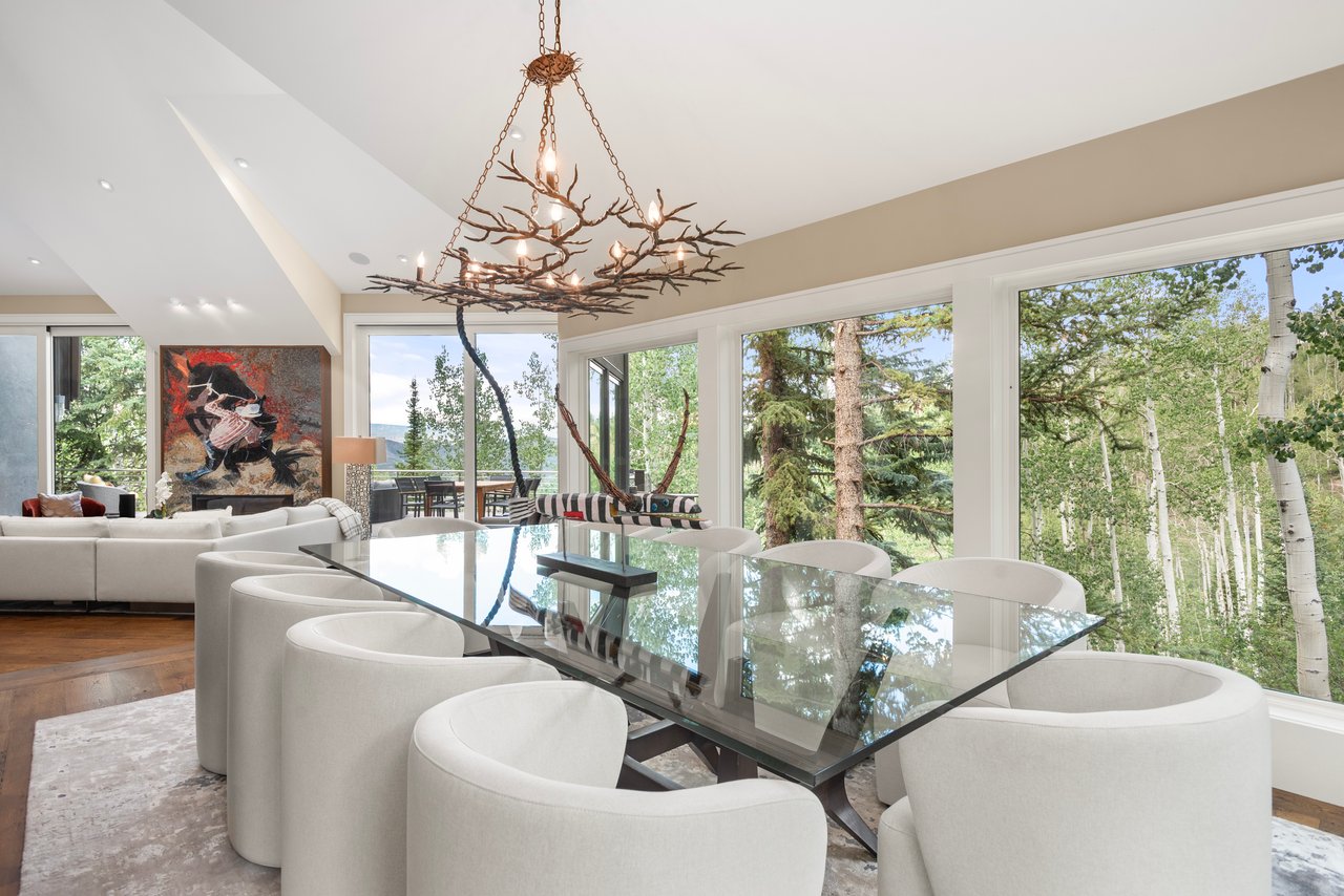 Dining room with a glass table, curved white chairs, branch chandelier, and large windows overlooking trees.