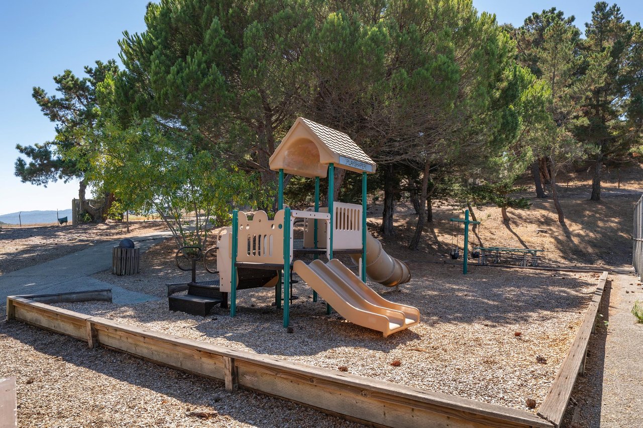 A children's playground with a slide and climbing structure set in a wood-chip play area in a sunny Belmont park.