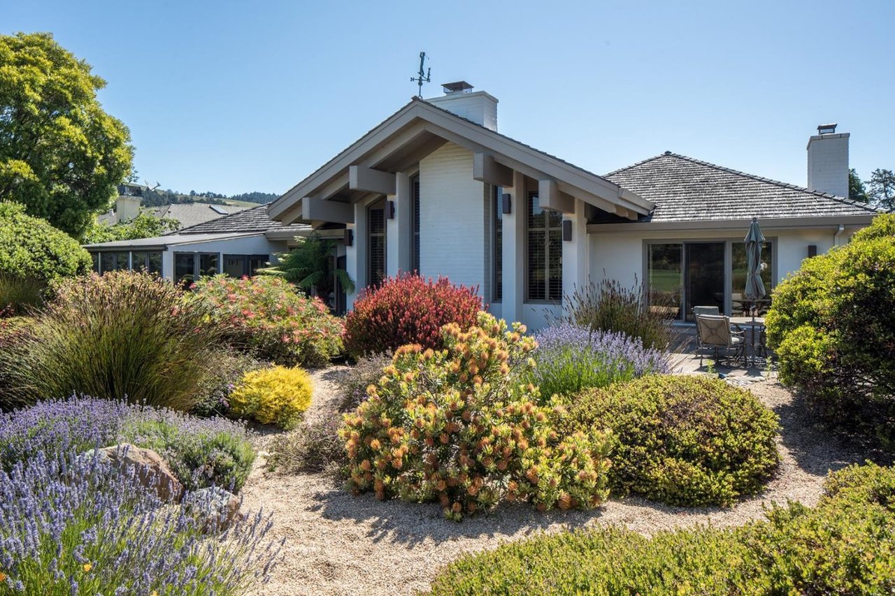 Single-story home with a gabled roof, flowering garden, gravel path, and patio seating.