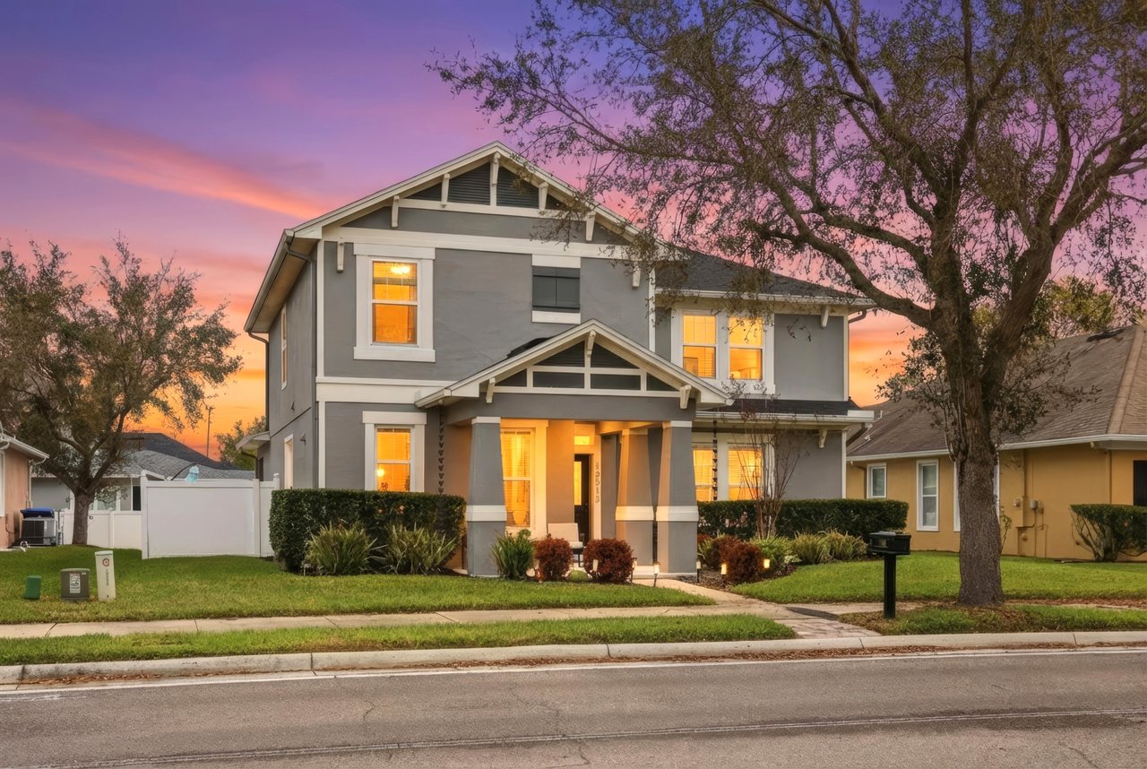 Two-story gray house with a covered porch and glowing windows beneath a pink sunset.