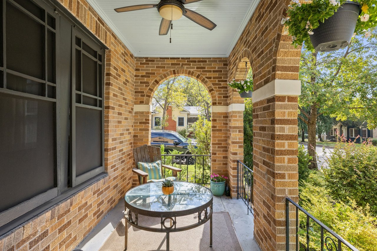 Covered brick porch with an arched opening, ceiling fan, hanging flowers, and round glass-top table.