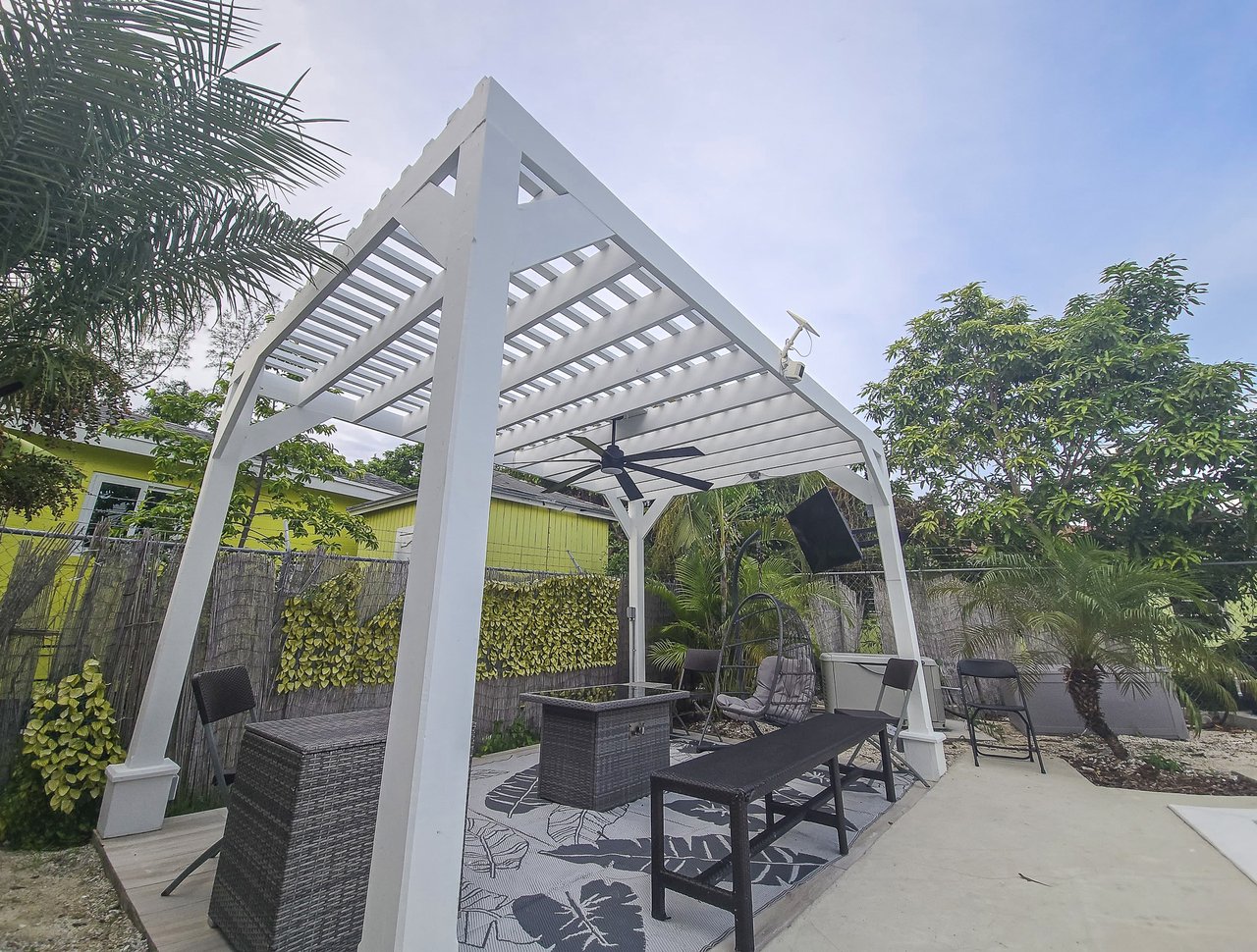 Furnished outdoor patio beneath a white pergola, with lounge seating and tropical landscaping.