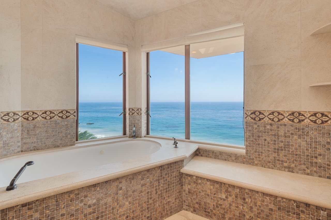 Built-in soaking tub beside large windows overlooking the ocean, with beige stone and patterned tile.