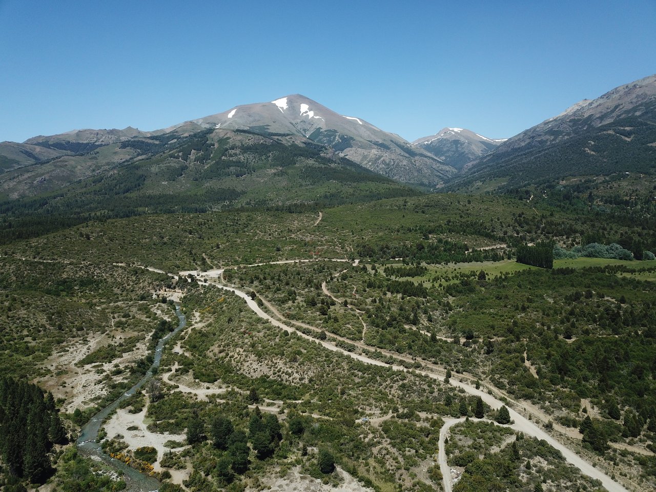 Aerial view of scrubland crossed by dirt roads and a stream, with snow-dusted mountains beyond.