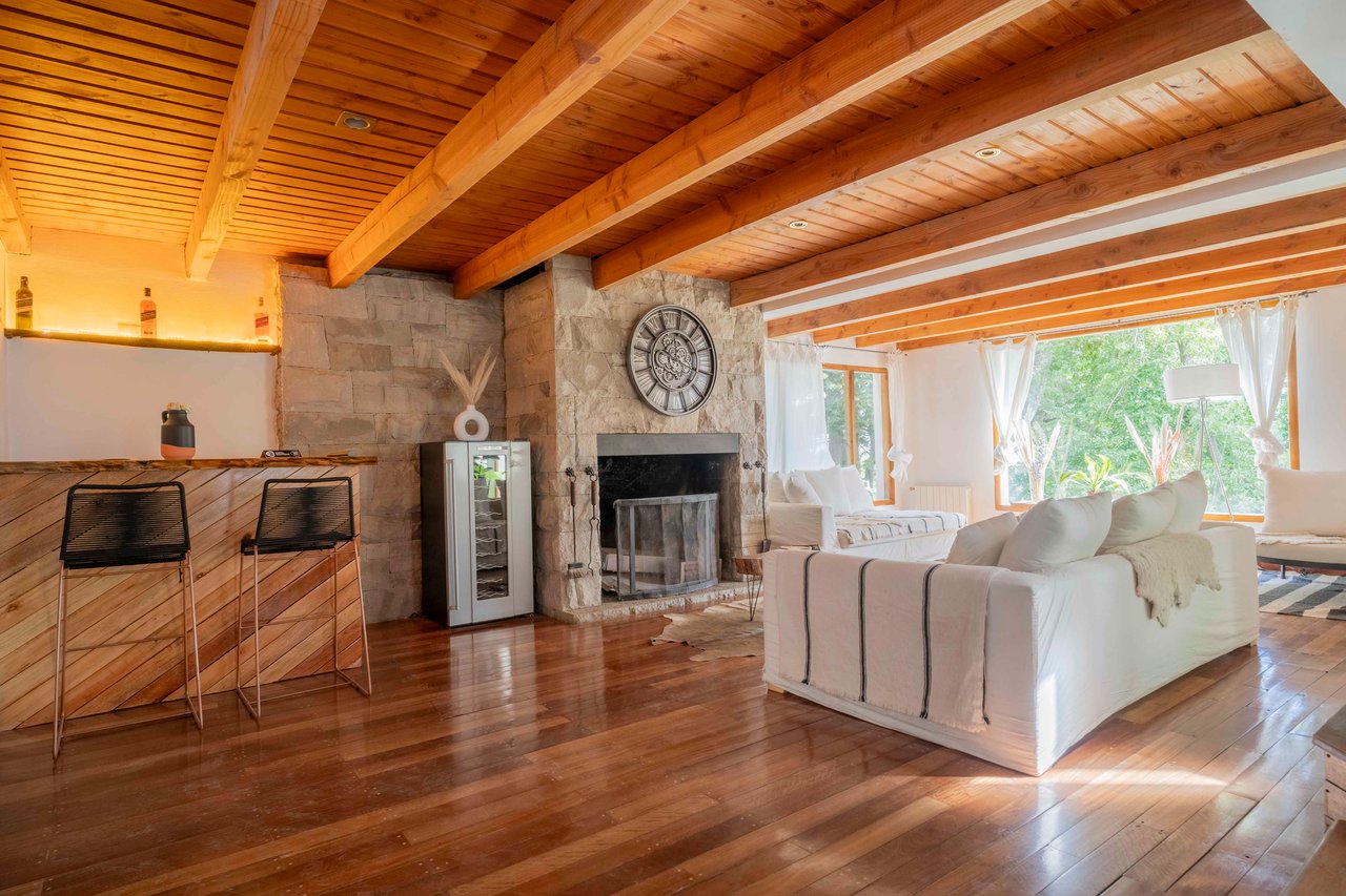 Living room with exposed timber beams, a stone fireplace, white sofas, and windows overlooking greenery.
