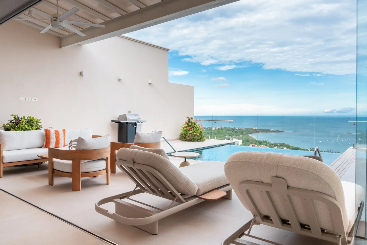 Covered condo terrace with poolside lounge chairs and an ocean view.