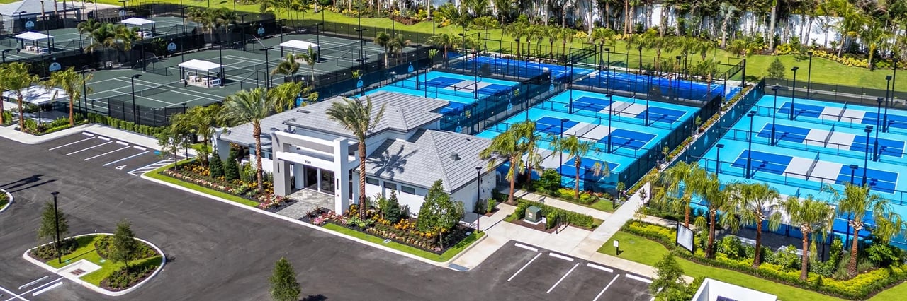 Aerial view of a clubhouse beside blue pickleball courts and tennis courts amid palm trees.