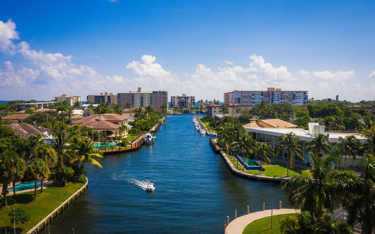 Aerial view of a waterfront neighborhood with a canal, palm-lined homes, docks, and distant condominiums.
