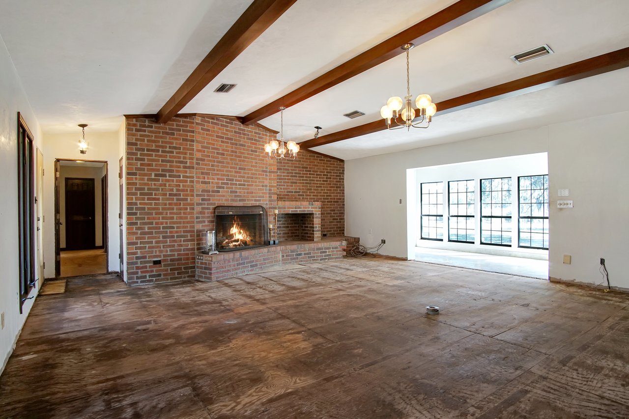 Spacious living room with exposed beams, brick fireplace, and warm light from chandeliers. Large windows overlook a sunny yard, creating an inviting ambiance.