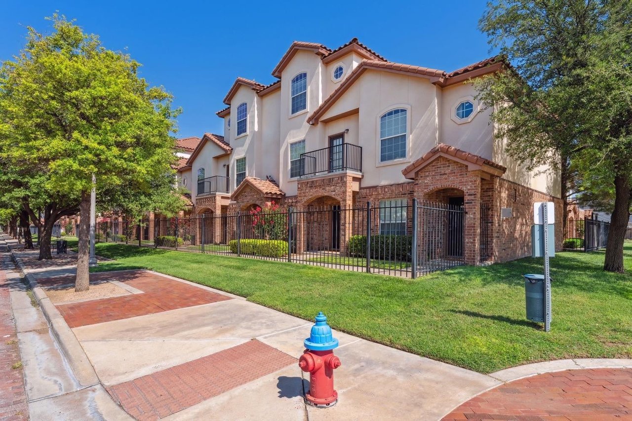 Attached two-story homes with brick and stucco exteriors, balconies, and a fenced lawn along a tree-lined sidewalk.