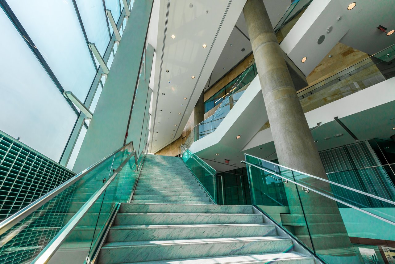 Modern atrium staircase with glass railings, tall windows, and a prominent concrete column.