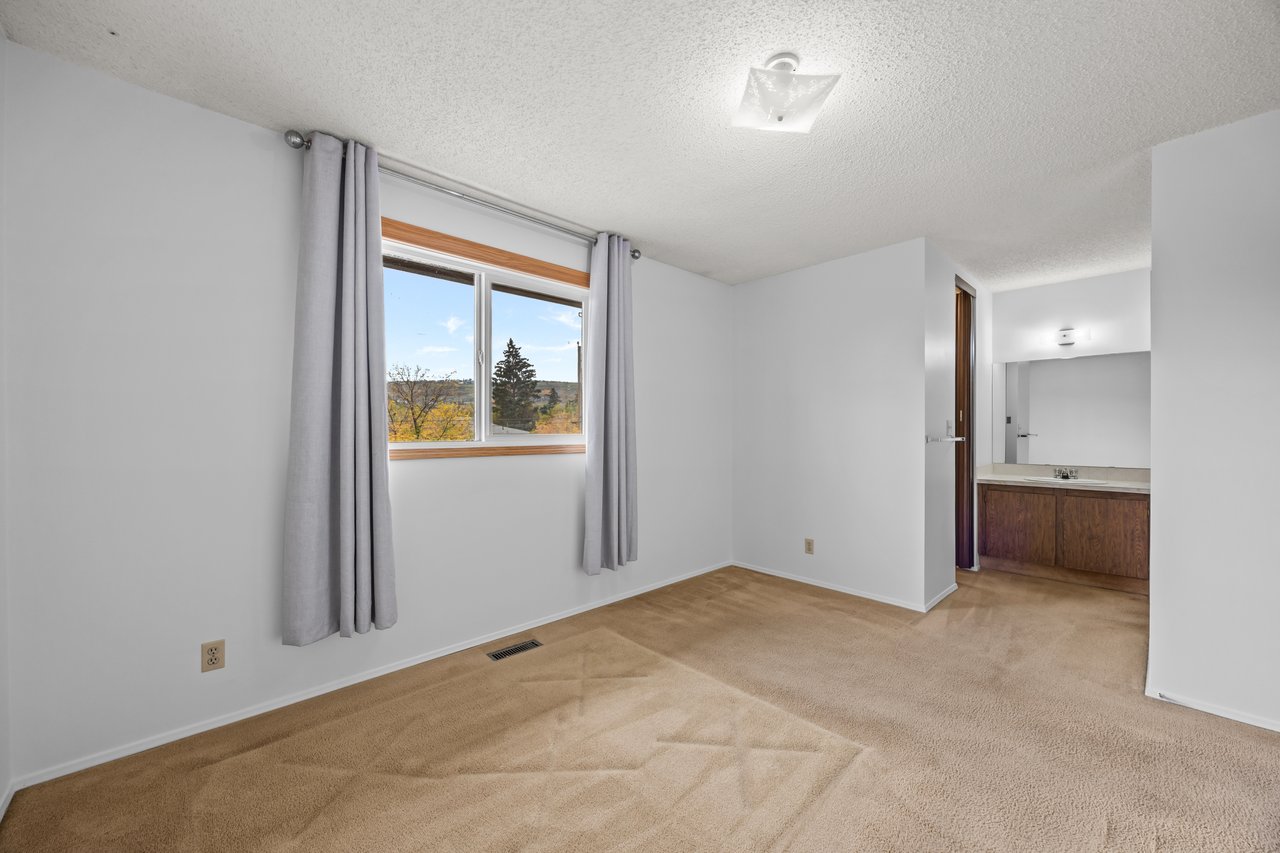 Empty carpeted bedroom with gray curtains, a window overlooking autumn trees, and an ensuite vanity.