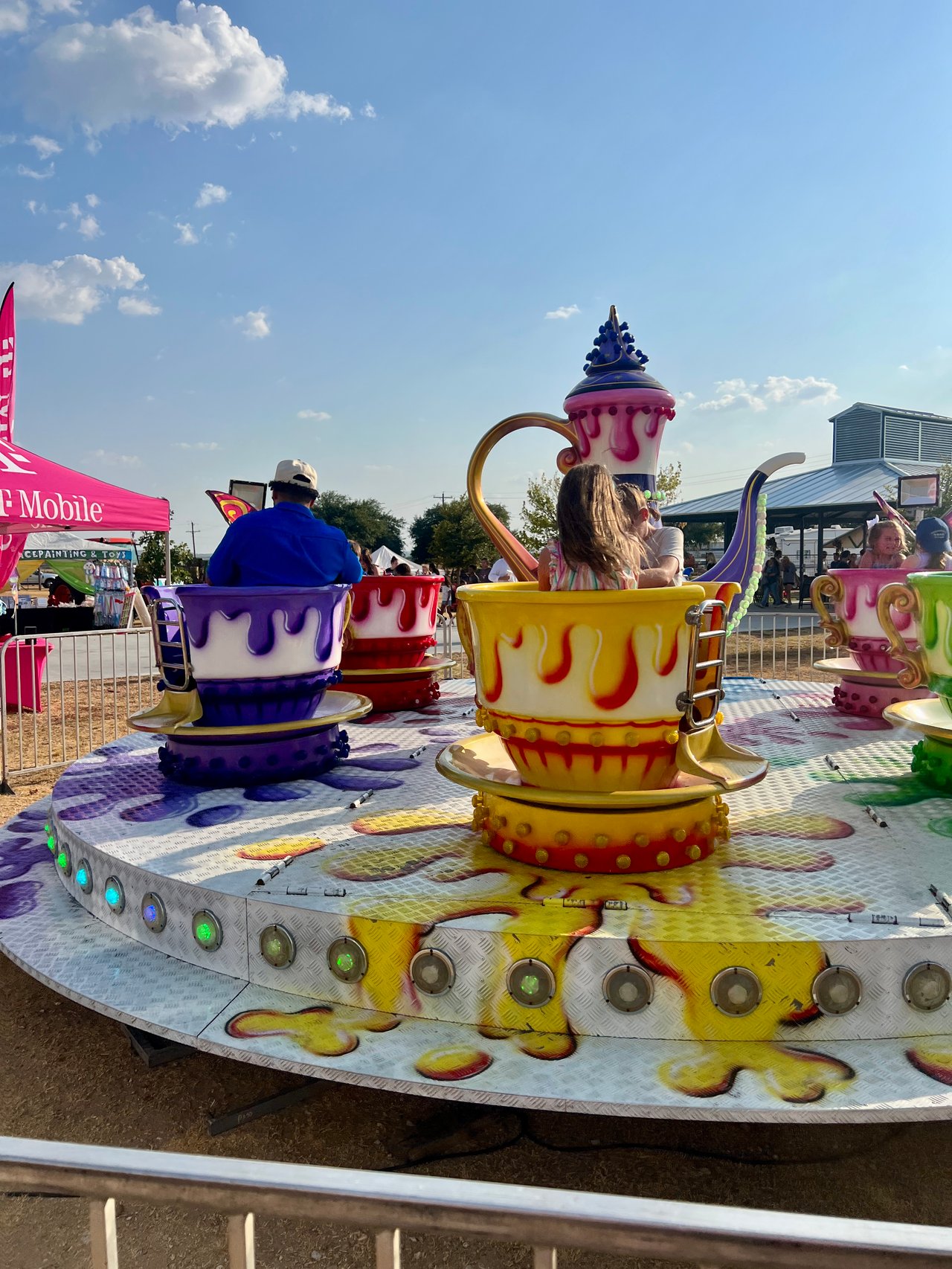 Fairgoers ride brightly colored teacup cars around a large teapot centerpiece at an outdoor fair.