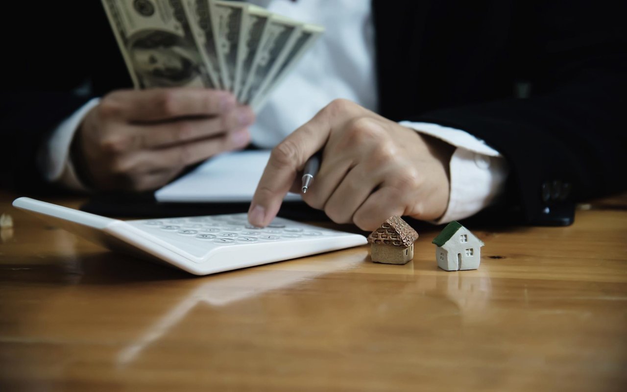 A suited person counts cash and uses a calculator beside two miniature houses.