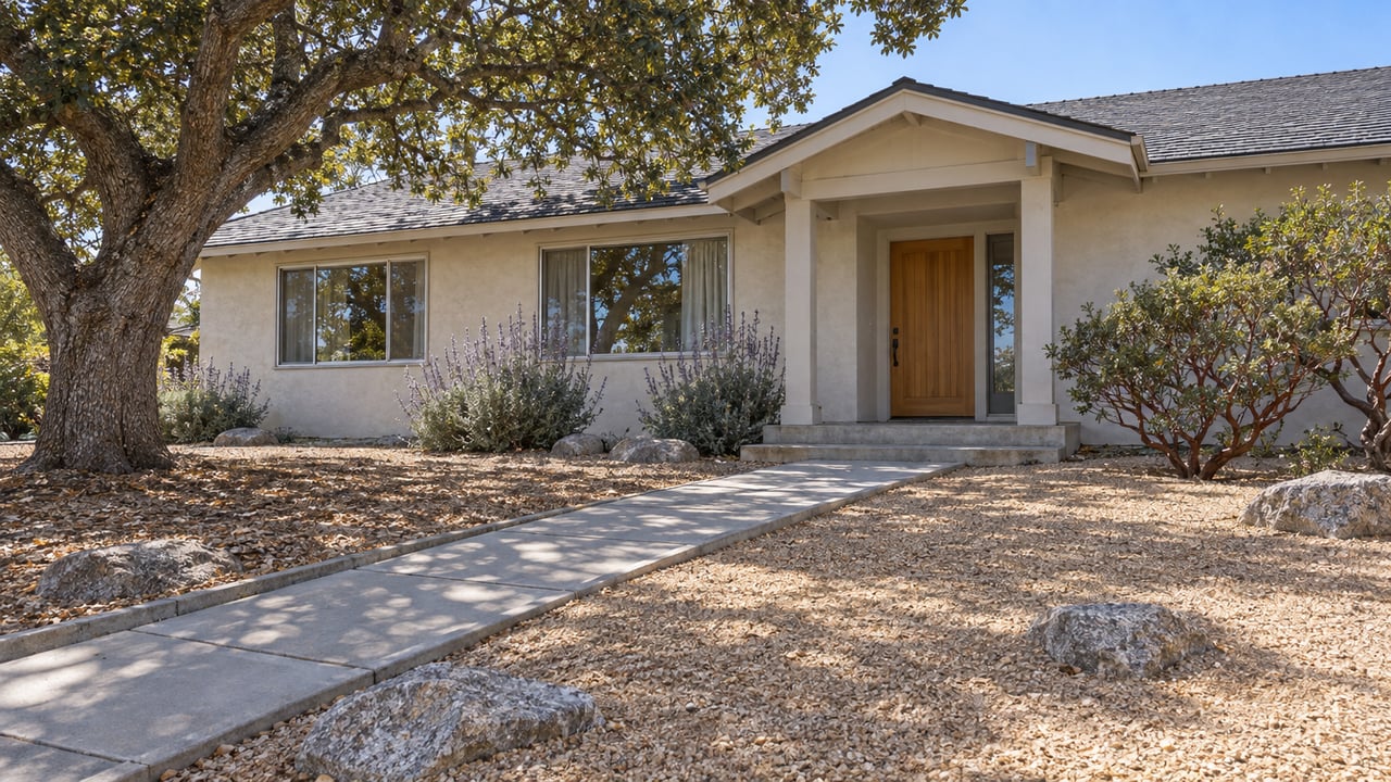 Low stucco ranch house behind a broad decomposed-granite yard, with a mature oak, flowering sage and a concrete front approach.