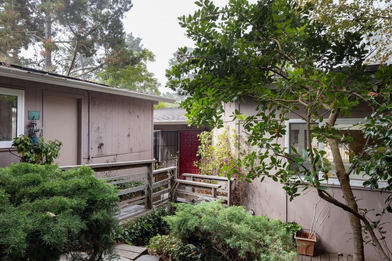 Home entrance with a red front door, wooden deck, and mature trees and shrubs.