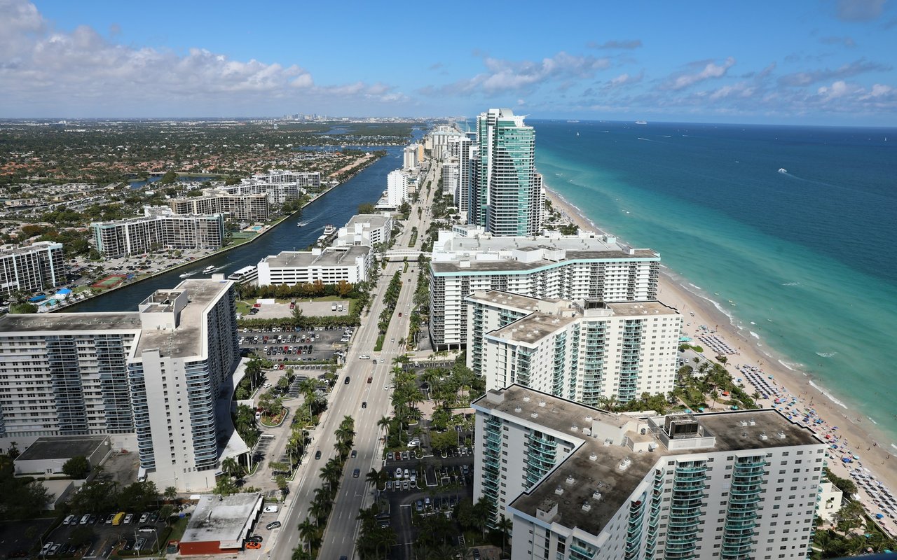 Coastal city high-rises beside a sandy beach, turquoise ocean, and palm-lined road.