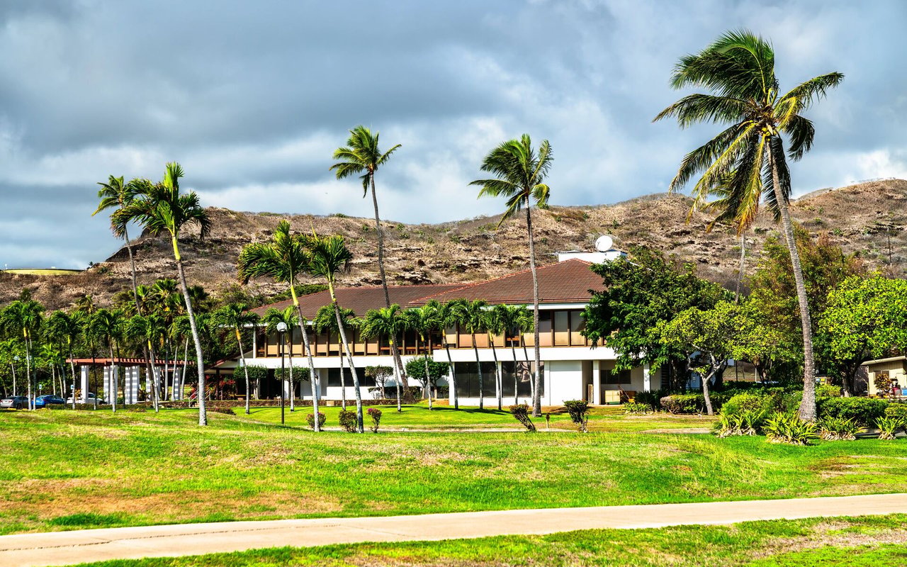 Low-rise building among palm trees, with a green lawn and dry hillside behind it.