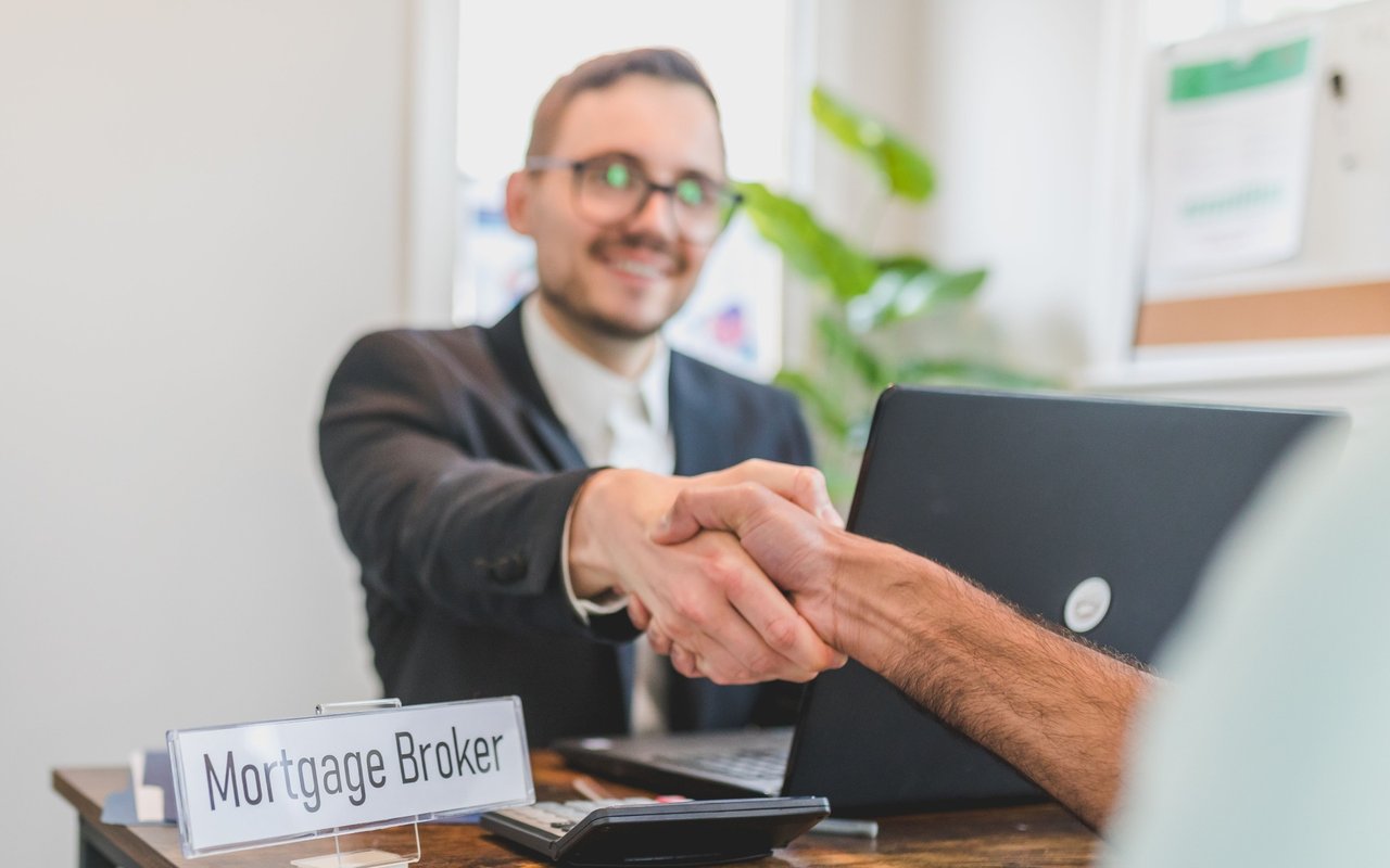 Customer shaking hands with a mortgage broker beside a desk sign reading “Mortgage Broker.”