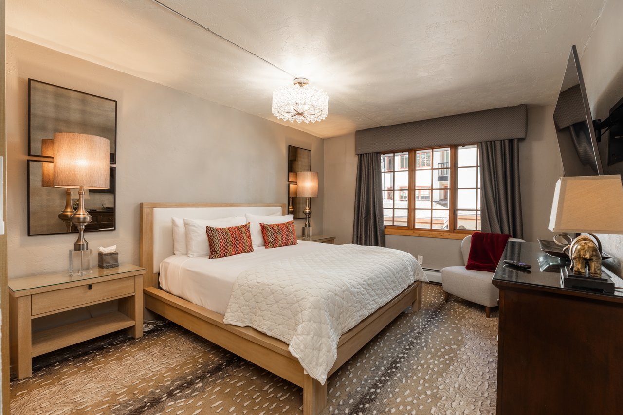Bedroom with a king-size bed, crystal chandelier, and window overlooking nearby buildings.