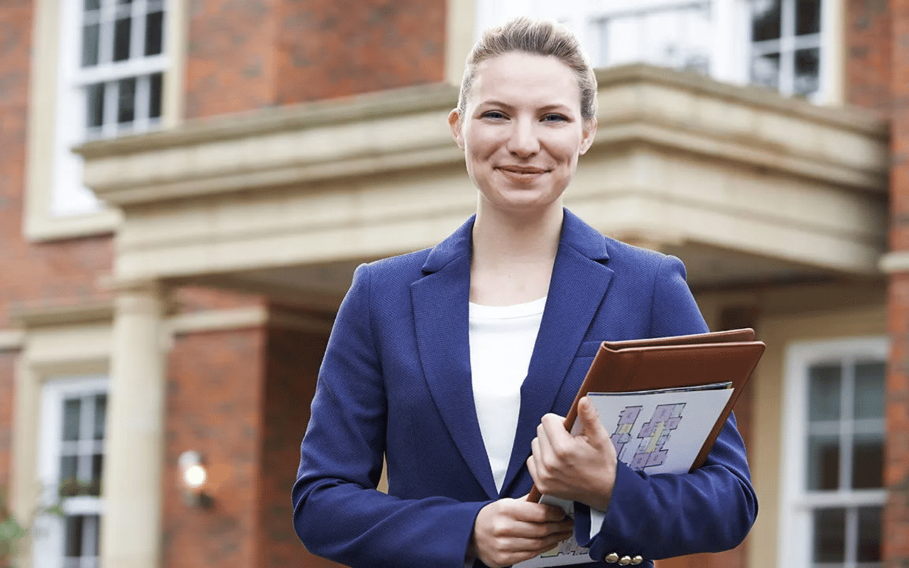 Real estate agent in a navy blazer holding a portfolio with floor plans outside a brick building.