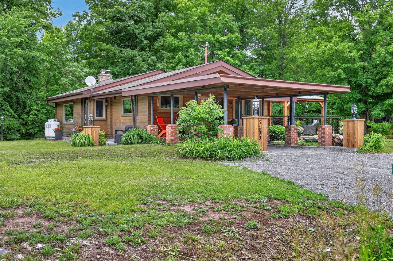 Wood-sided cottage with a broad covered porch, brick supports, and gravel driveway amid leafy trees.