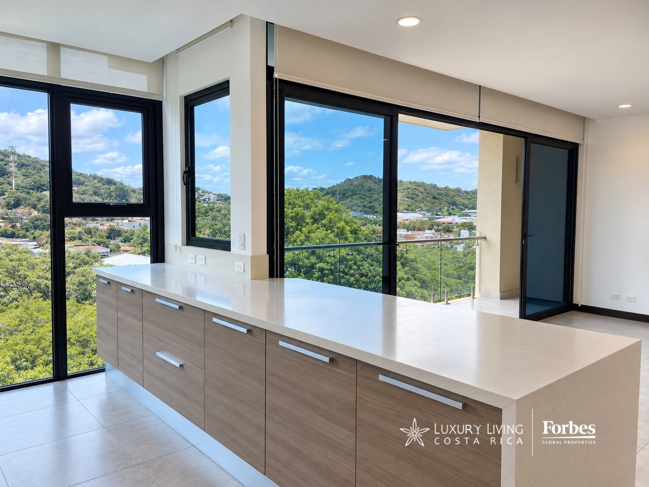Modern kitchen with wood-front cabinets, a long white counter, and windows overlooking green hills.