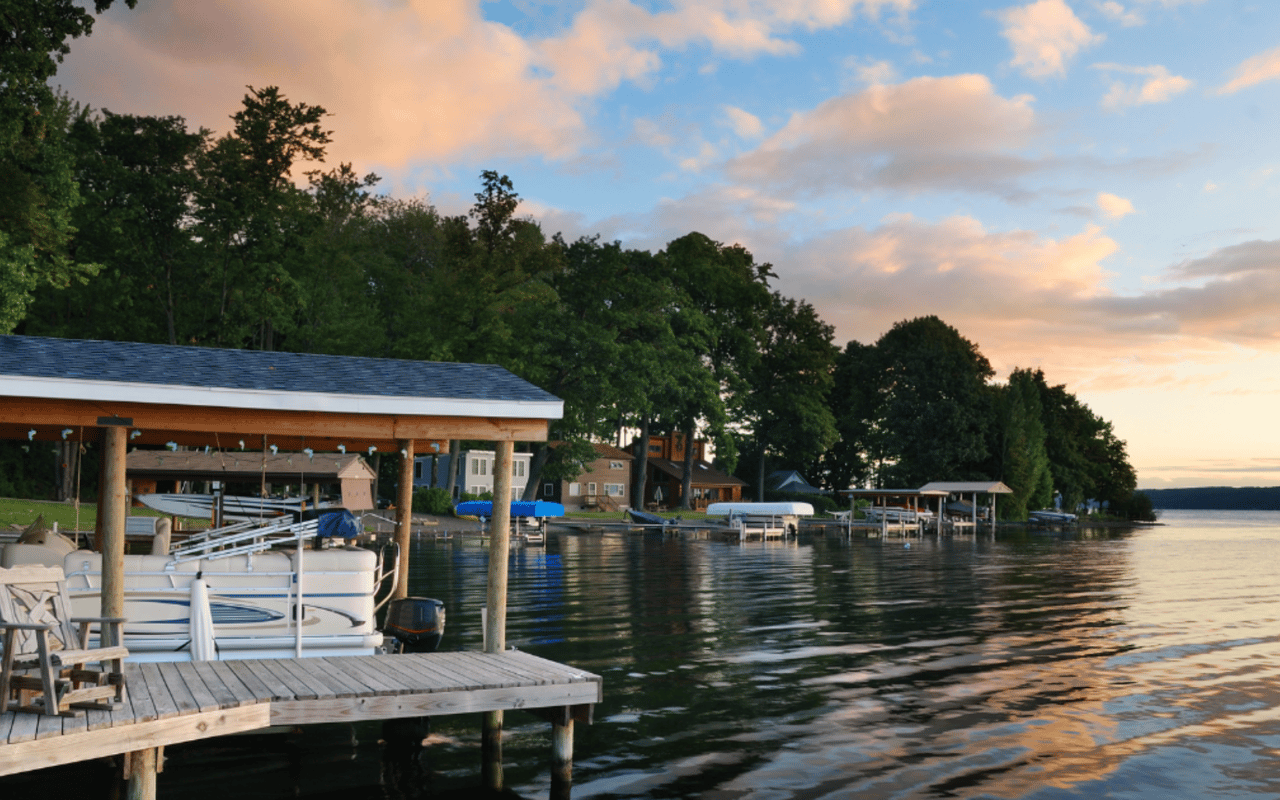 Lakefront homes and covered docks beside calm water at sunset, with a pontoon boat in the foreground.