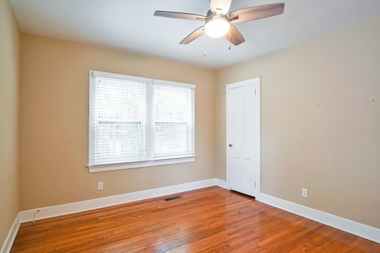 Empty room with light beige walls, a ceiling fan, and hardwood floor. Two large windows with blinds and a white door enhance the bright, spacious feel.