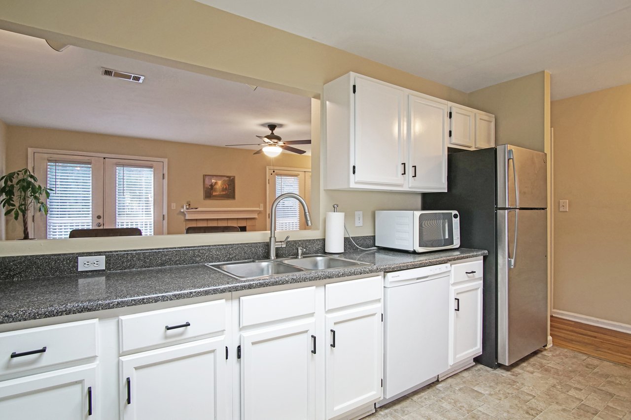 Kitchen at 529 Collinsford Road, Tallahassee, Florida, featuring white cabinetry with black hardware, gray countertops, and stainless steel appliances including a refrigerator and dishwasher. The kitchen opens to the living room through a pass-through window with views of the fireplace and French doors leading to the back deck.