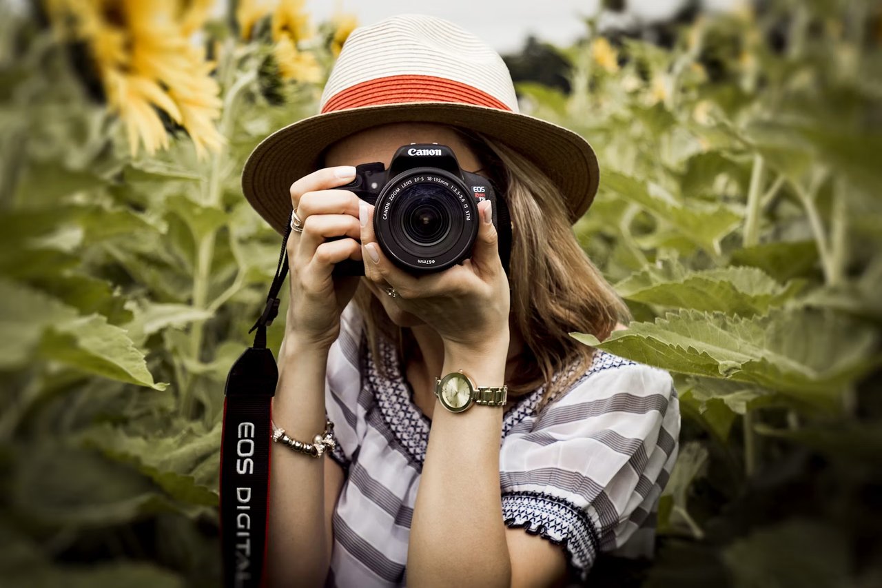 A woman in a sun hat photographs through a Canon camera among leafy plants.
